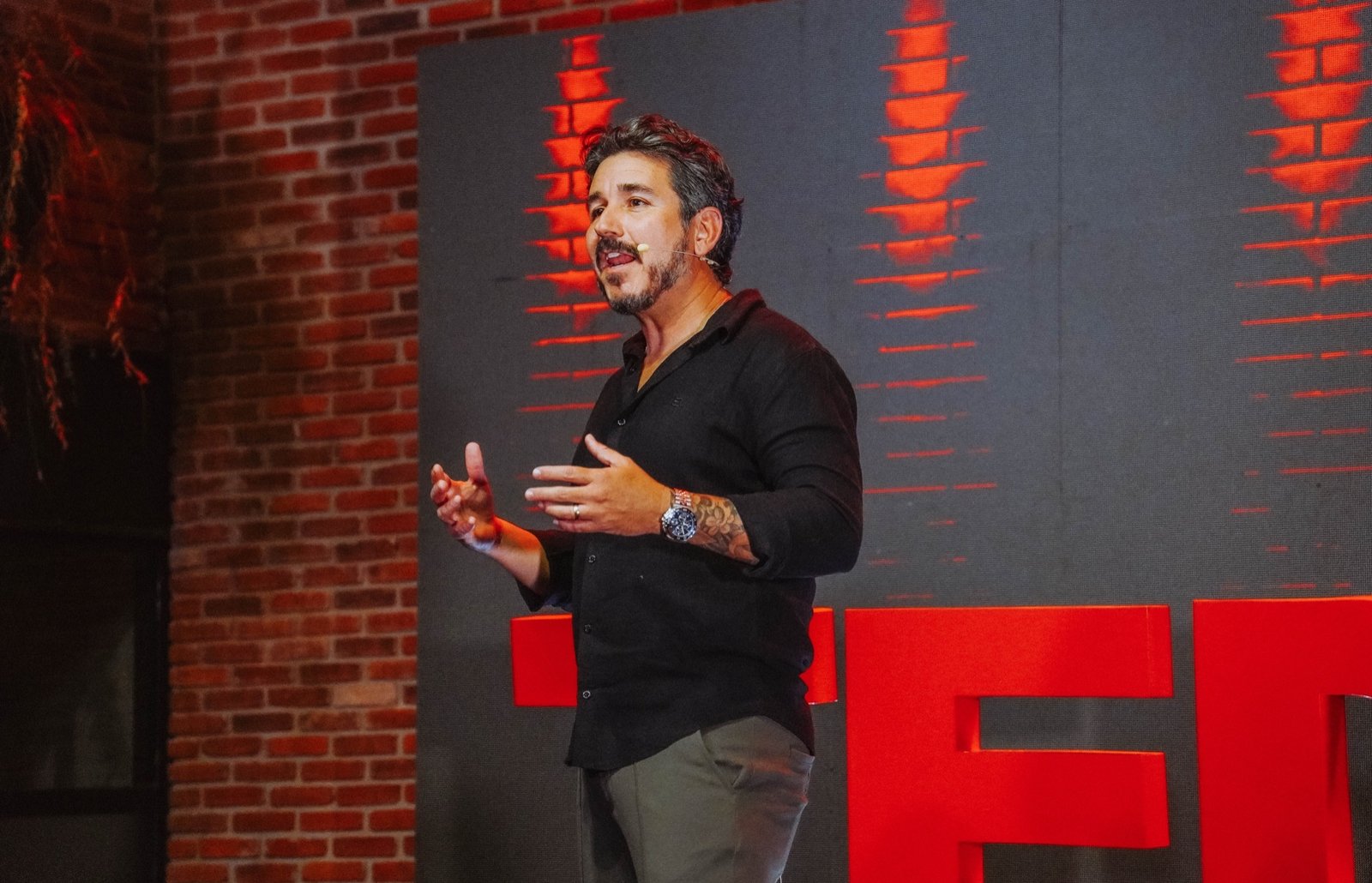 Retrato de Dan Rocha no palco, microfone de lapela, luz vermelha ao fundo
